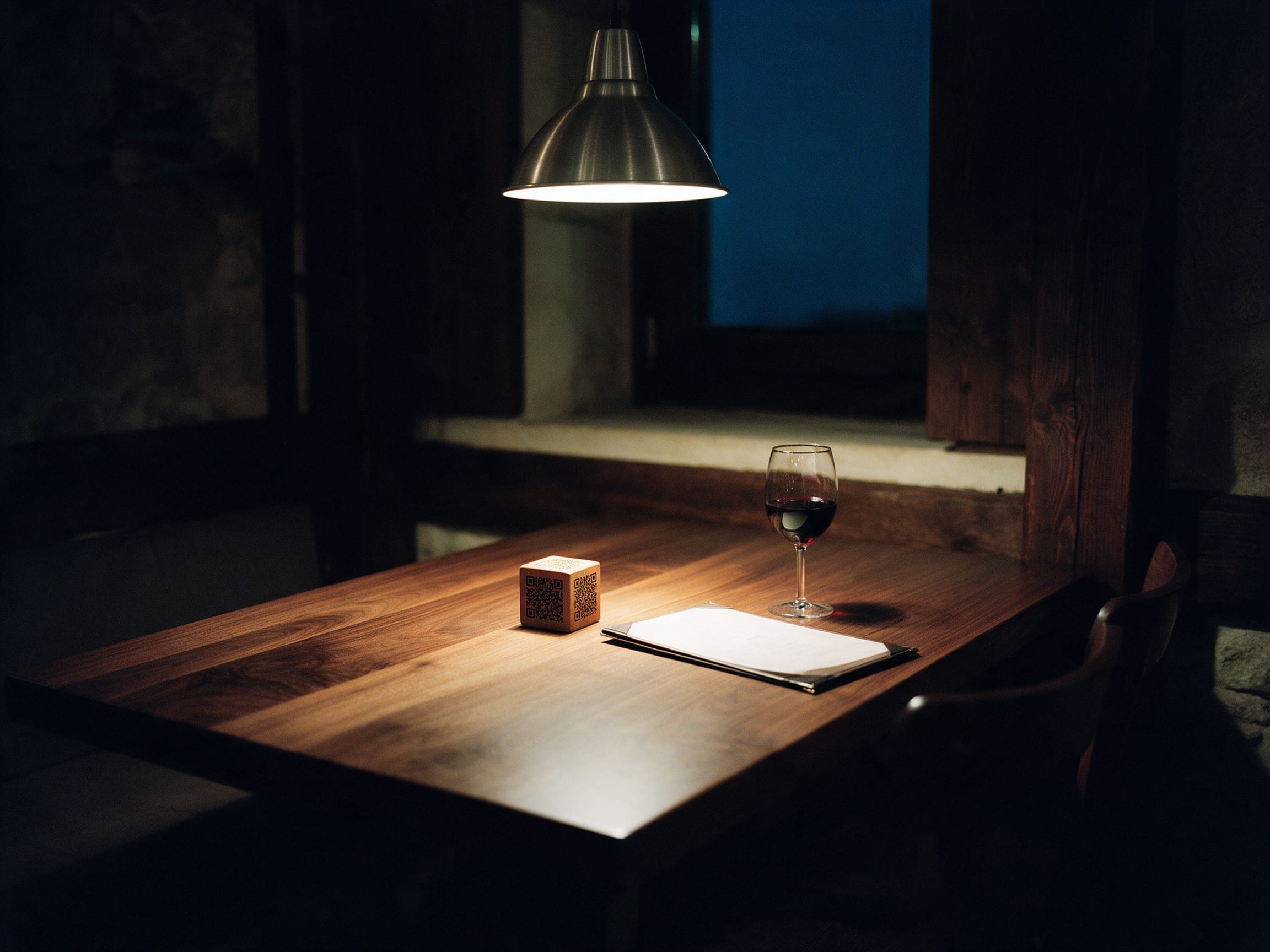 A laid restaurant table at night with a wooden QR cube under a pendant lamp