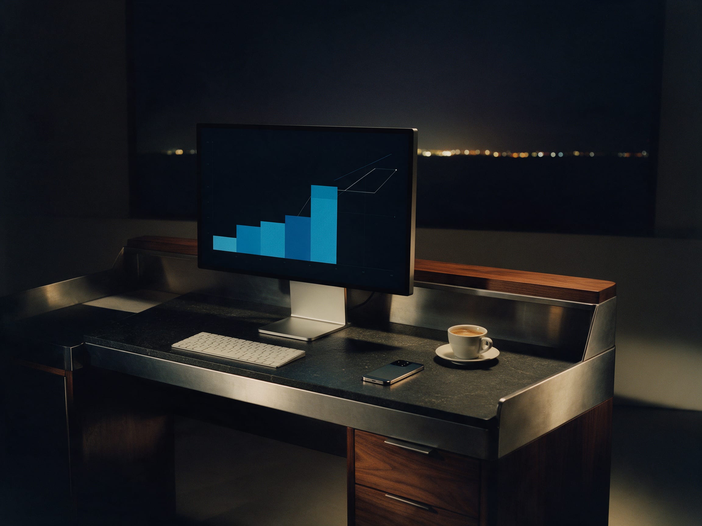 A monitor showing abstract rising charts on a dark desk at night, a phone and coffee beside it